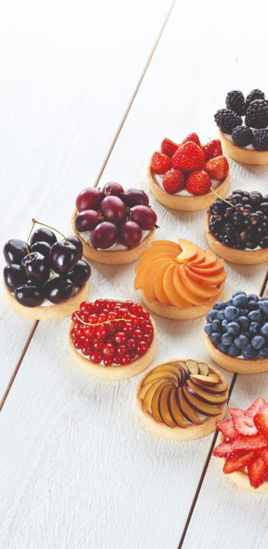 Kleine Tartelettes gefüllt mit verschiedenen Beeren und Obstsorten auf einem Holztisch.